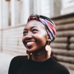 Smiling woman wearing vibrant headscarf and earrings, captured in an urban street setting.