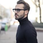 A fashionable man in a black top and glasses poses in an urban street with parked cars.